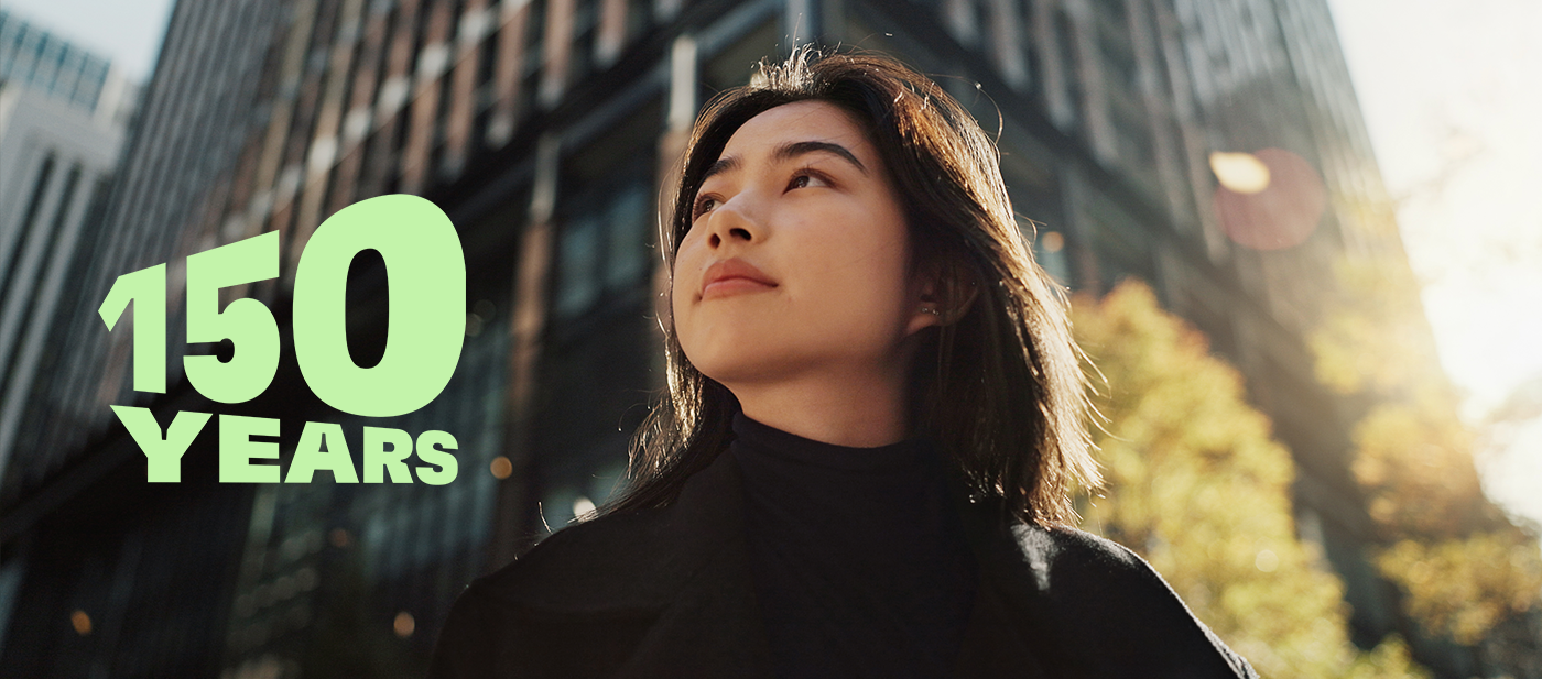 Texte vert indiquant « 150 ANS » sur un paysage urbain moderne avec de grands bâtiments en verre et des arbres d'automne, symbolisant la célébration du 150e anniversaire.