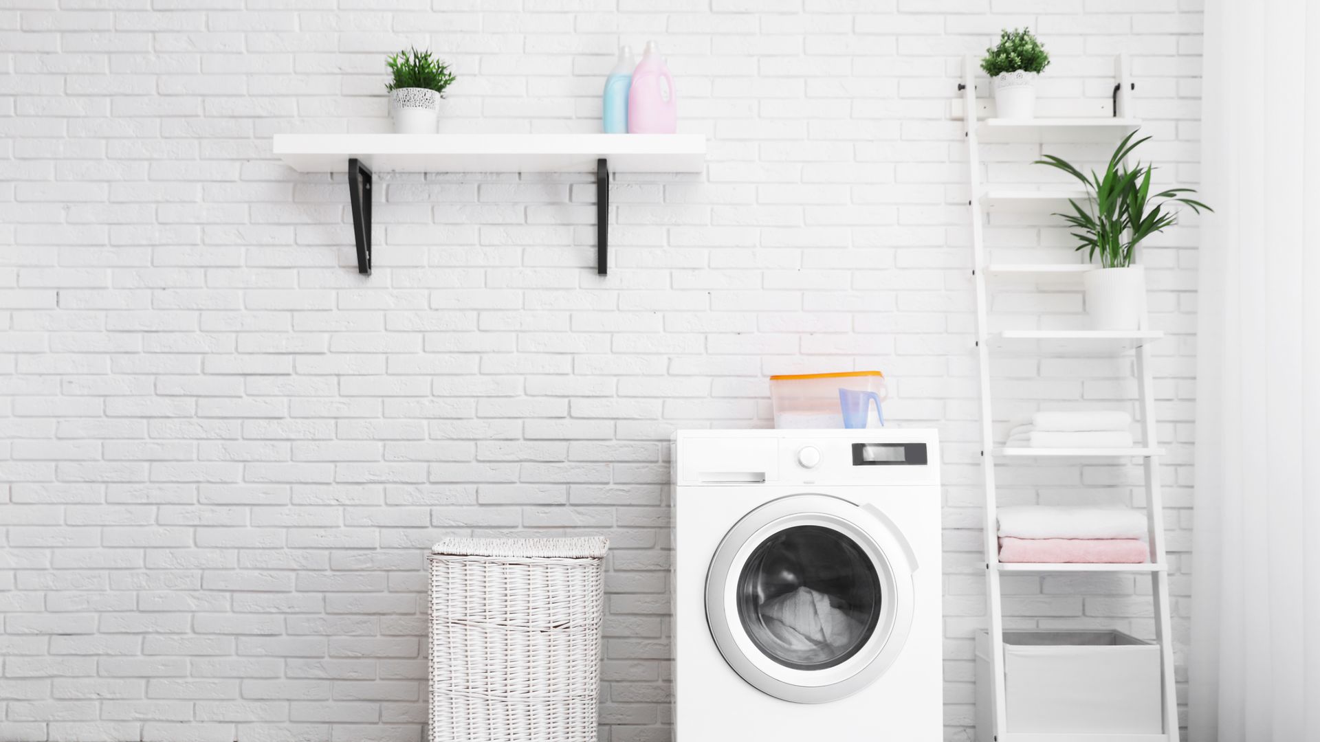A bathroom with a washing machine, a laundry basket and two shelves with plants on them