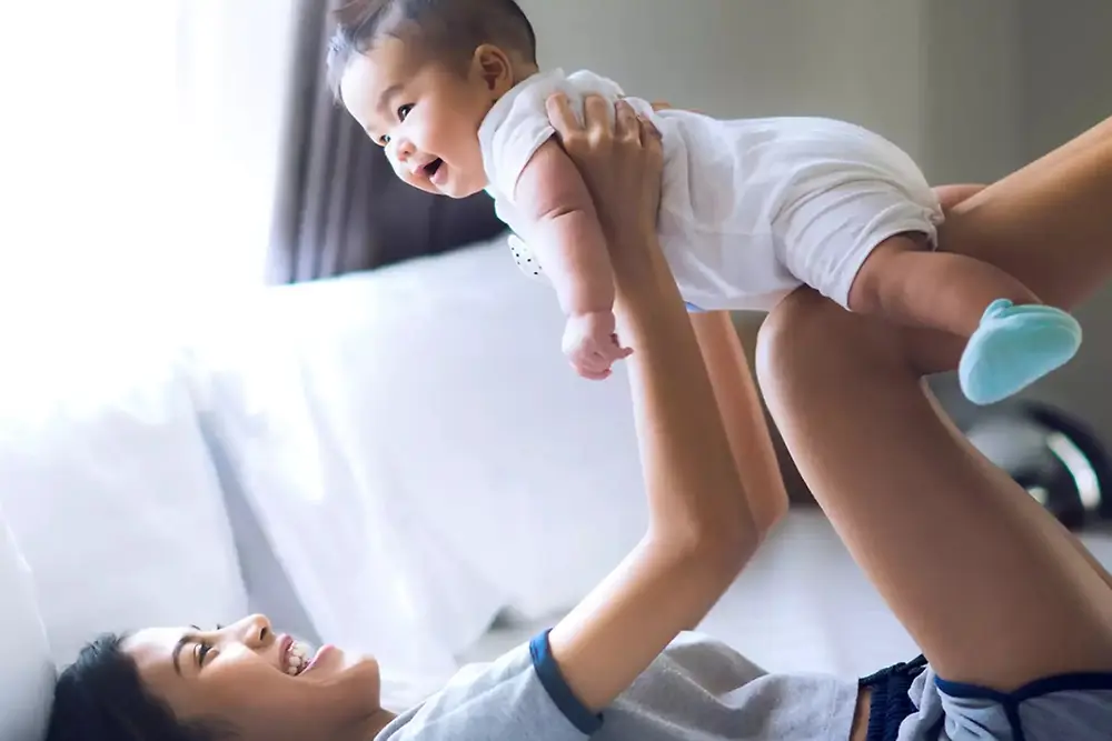 Une mère soulève et joue avec un bébé souriant à l'intérieur, dans une maison lumineuse.