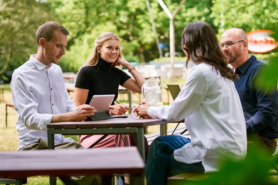 Groupe de collègues assis ensemble à l'extérieur, discutant du travail à l'aide de tablettes et d'ordinateurs portables, illustrant la collaboration et une culture ouverte, tournée vers l'esprit d'équipe, chez Henkel.