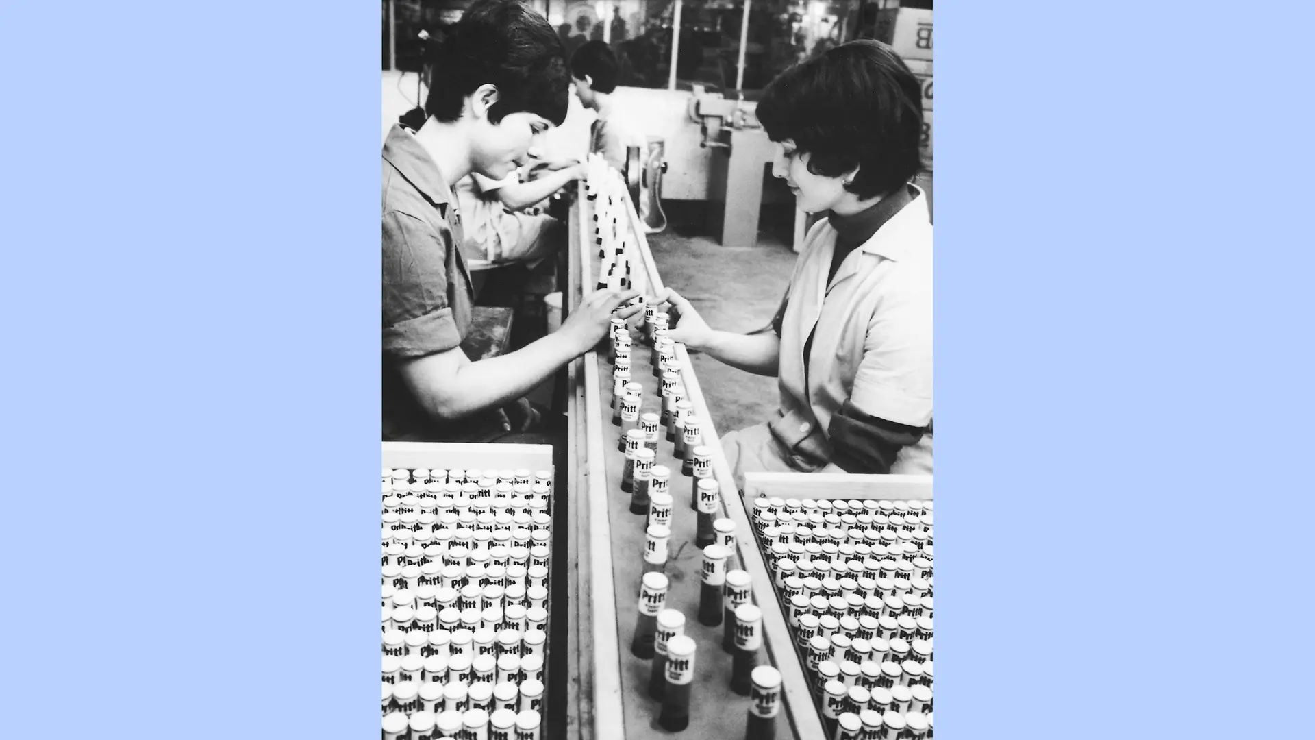 photo en noir et blanc de l’usine de conditionnement des produits Pritt. Deux femmes sont assises face à face de part et d’autre d’un tapis roulant sur lequel des sticks de colle Pritt sont disposés en rangées. À côté de chaque femme se trouve une boîte remplie de sticks Pritt supplémentaires.