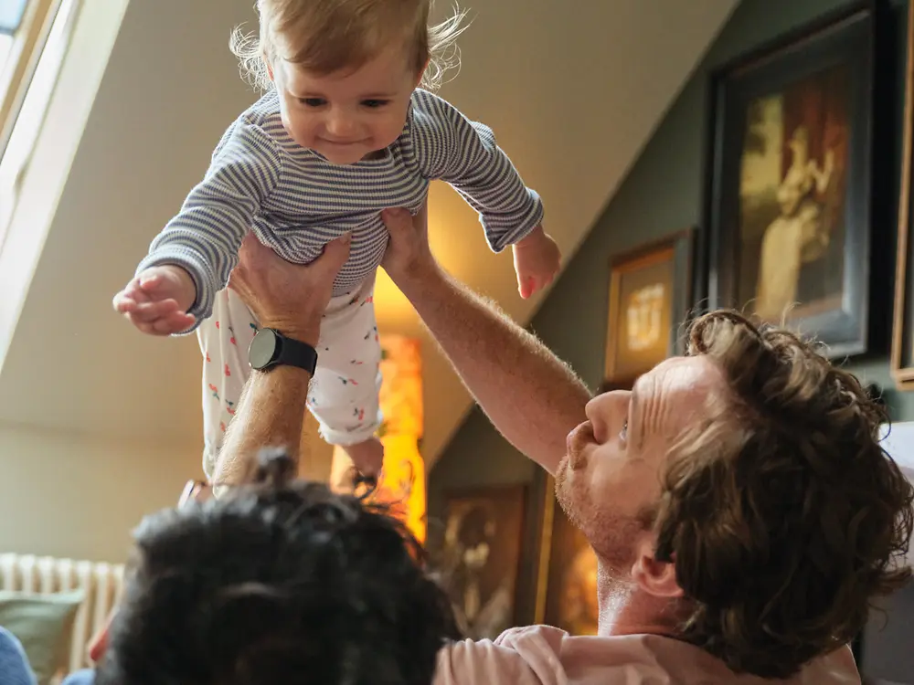 Deux pères tenant leur bébé et souriant à la maison, symbolisant l’inclusivité et la politique de congé parental favorable à la famille de Henkel.