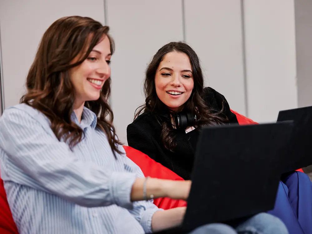 Deux personnes assises sur des fauteuils poires colorés dans un bureau moderne, chacune utilisant un ordinateur portable. Elles semblent discuter des avantages sociaux de Henkel tout en travaillant en collaboration.