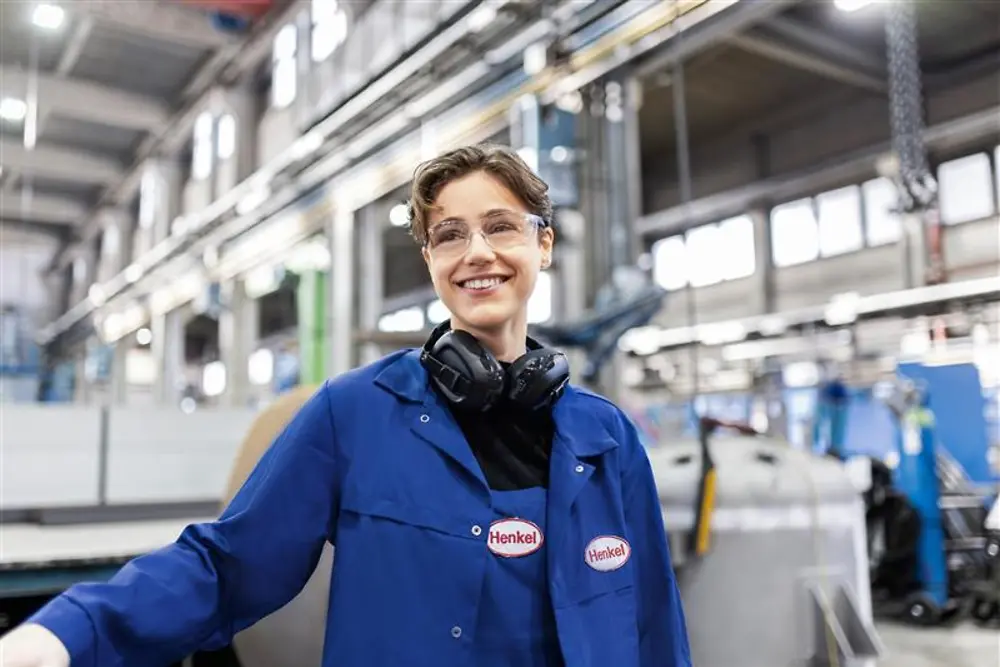 Un membre du personnel de Henkel souriant, portant une blouse de travail bleue et un casque de sécurité, se tenant dans un hall de production industrielle, représentant l'expertise et l'innovation dans la fabrication.