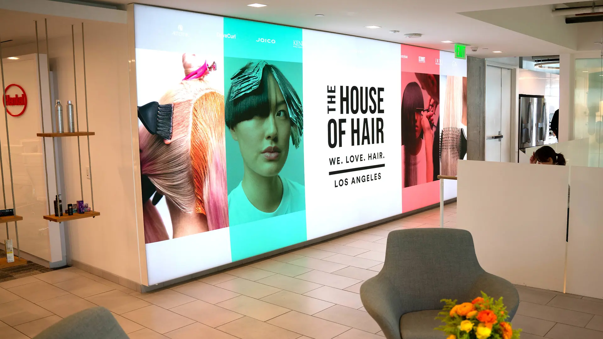 Un hall moderne avec un mur lumineux affichant de grands panneaux graphiques colorés sur le thème des cheveux et le texte « THE HOUSE OF HAIR – WE. LOVE. HAIR. – LOS ANGELES ». L’espace comprend des fauteuils lounge gris clair, une petite table avec un bouquet de fleurs vif, et un design épuré avec sol carrelé et cloisons blanches.