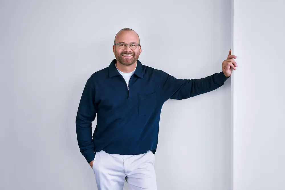 Professionnel expérimenté chez Henkel, debout, confiant, contre un mur blanc dans un cadre moderne, vêtu d'une chemise foncée à manches longues et d'un pantalon de couleur claire.
