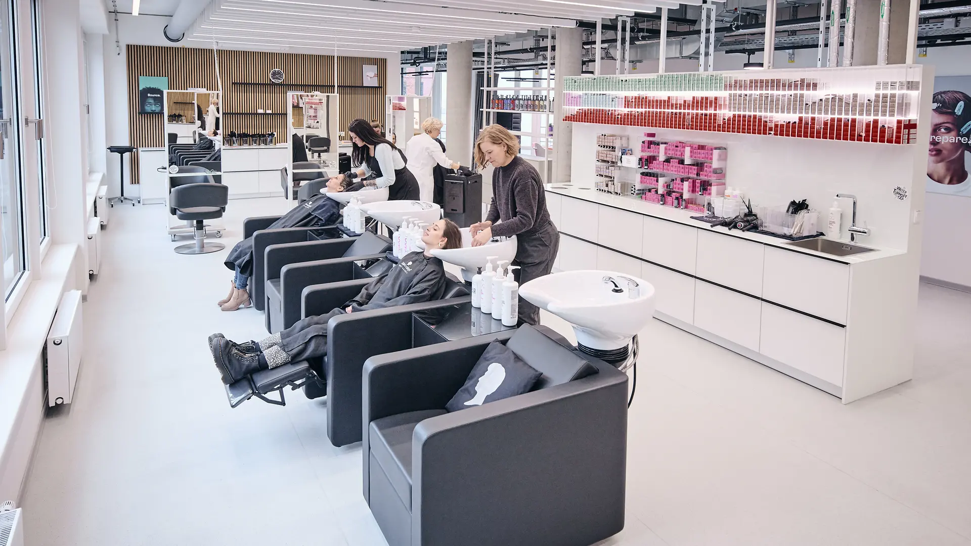 Salon de coiffure avec plusieurs bacs à shampooing. Des clients test sont assis sur deux fauteuils, leurs cheveux étant lavés par deux employés.