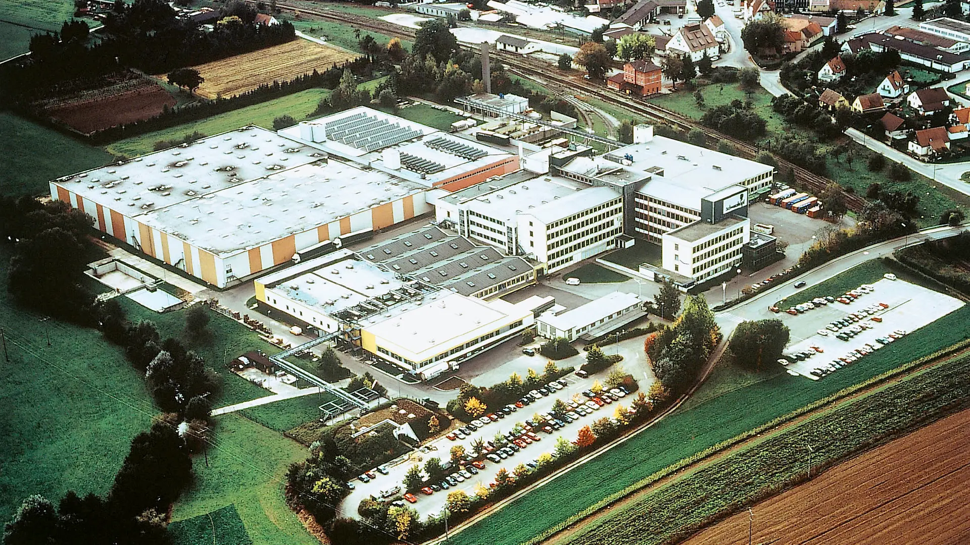 Une vue aérienne de l’usine Schwarzkopf à Wassertrüdingen en 1995, montrant plusieurs bâtiments et halls de production, ainsi qu’un grand parking rempli de voitures.
