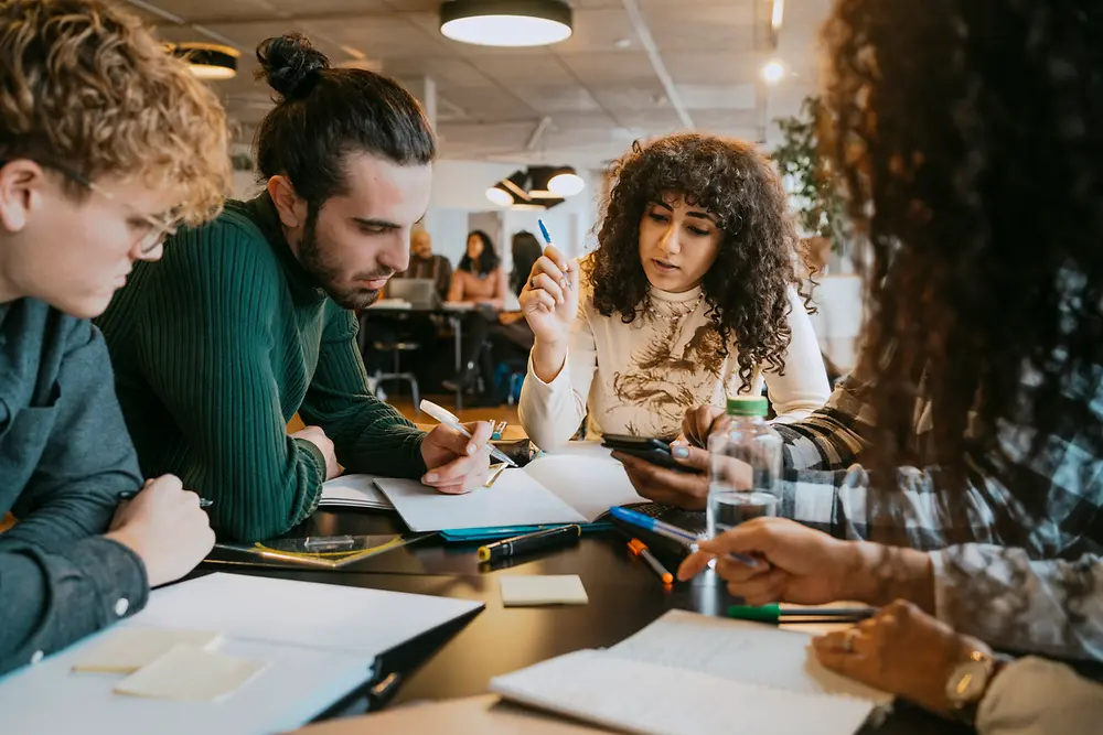 Un groupe de personnes assises autour d’une table dans un espace de travail intérieur moderne, collaborant sur des documents, des carnets et des outils numériques. Divers stylos, feuilles de papier et notes autocollantes sont disposés sur la table, tandis que le groupe échange et prend des notes.