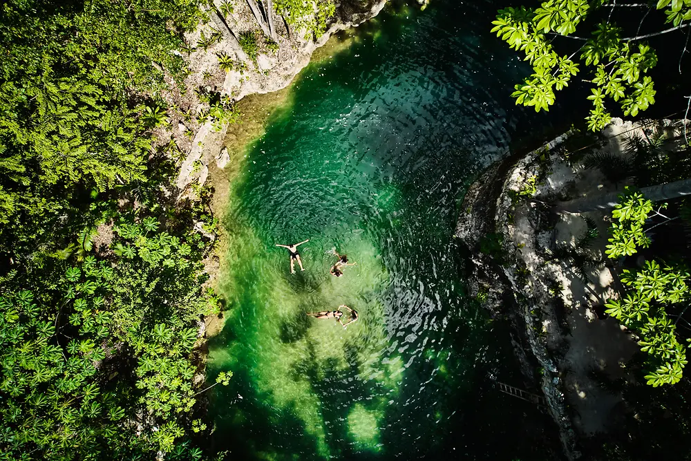 Vue aérienne d’un petit bassin naturel entouré d’une végétation dense et verdoyante, avec trois personnes flottant dans une eau claire aux reflets émeraude.
