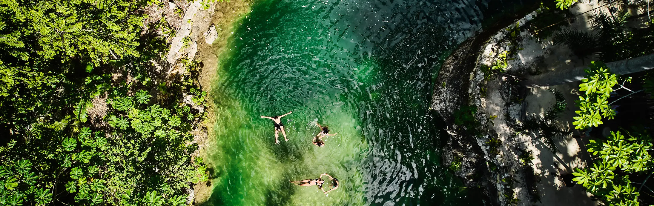 Vue aérienne d’un petit bassin naturel entouré d’une végétation dense et verdoyante, avec trois personnes flottant dans une eau claire aux reflets émeraude.