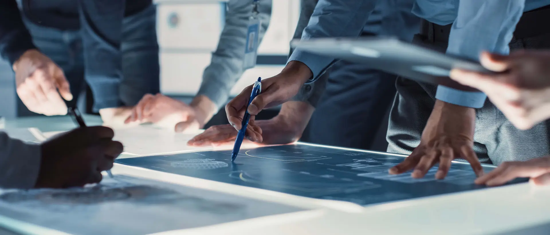 Vue rapprochée de plusieurs personnes réunies autour d’une grande table lumineuse, examinant et annotant des plans techniques à l’aide de stylos et d’outils numériques.