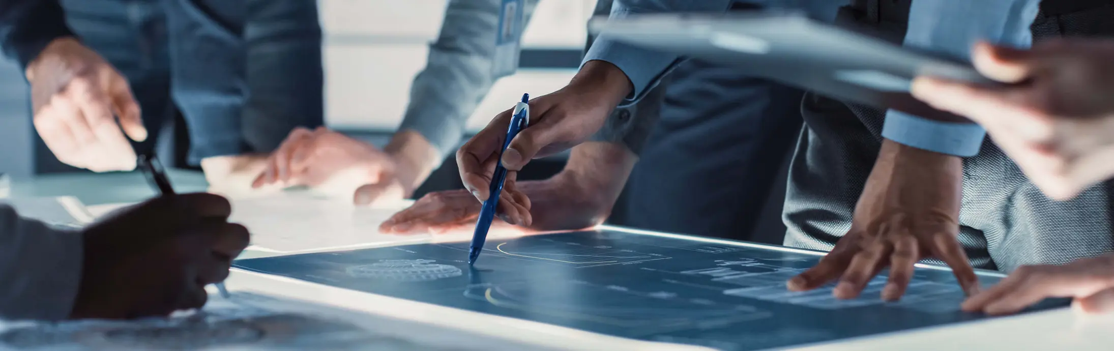 Vue rapprochée de plusieurs personnes réunies autour d’une grande table lumineuse, examinant et annotant des plans techniques à l’aide de stylos et d’outils numériques.