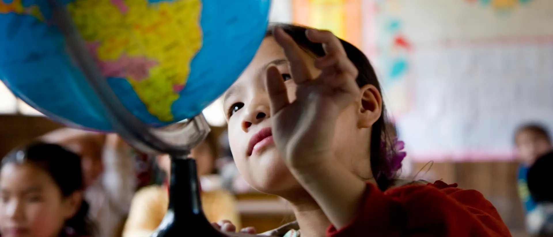 Un enfant dans une salle de classe tend la main vers un grand globe posé sur un bureau, tandis que des cartes colorées et des affiches sont visibles sur les murs en arrière‑plan.