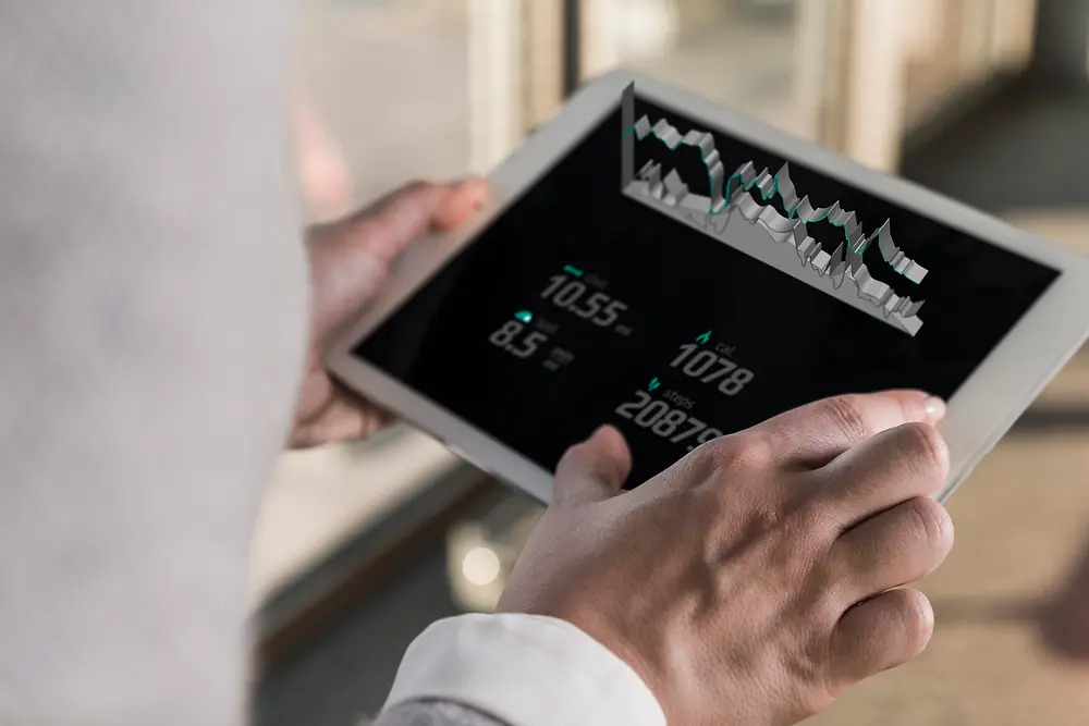 Une personne tenant une tablette affichant des graphiques numériques et des données chiffrées, vue par‑dessus l’épaule dans un environnement intérieur lumineux.